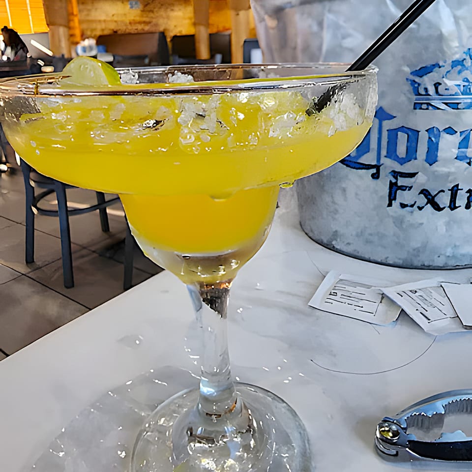 A vibrant yellow margarita with a salted rim and lime wedge, next to a Corona Extra ice bucket.
