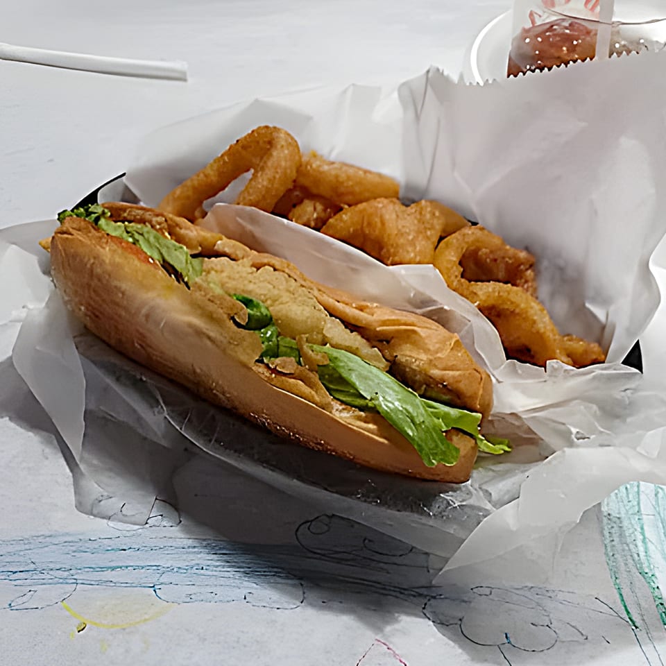 A fried seafood po'boy sandwich with lettuce and a side of onion rings in a paper-lined basket.