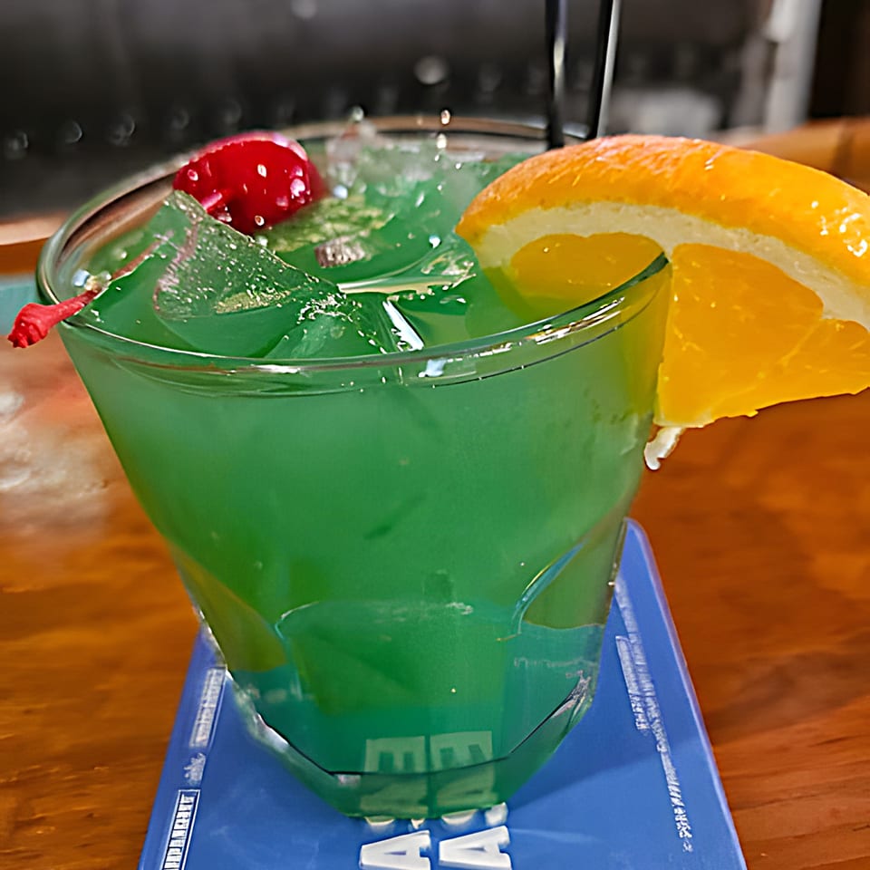 Green cocktail with ice, garnished with a maraschino cherry and an orange slice.